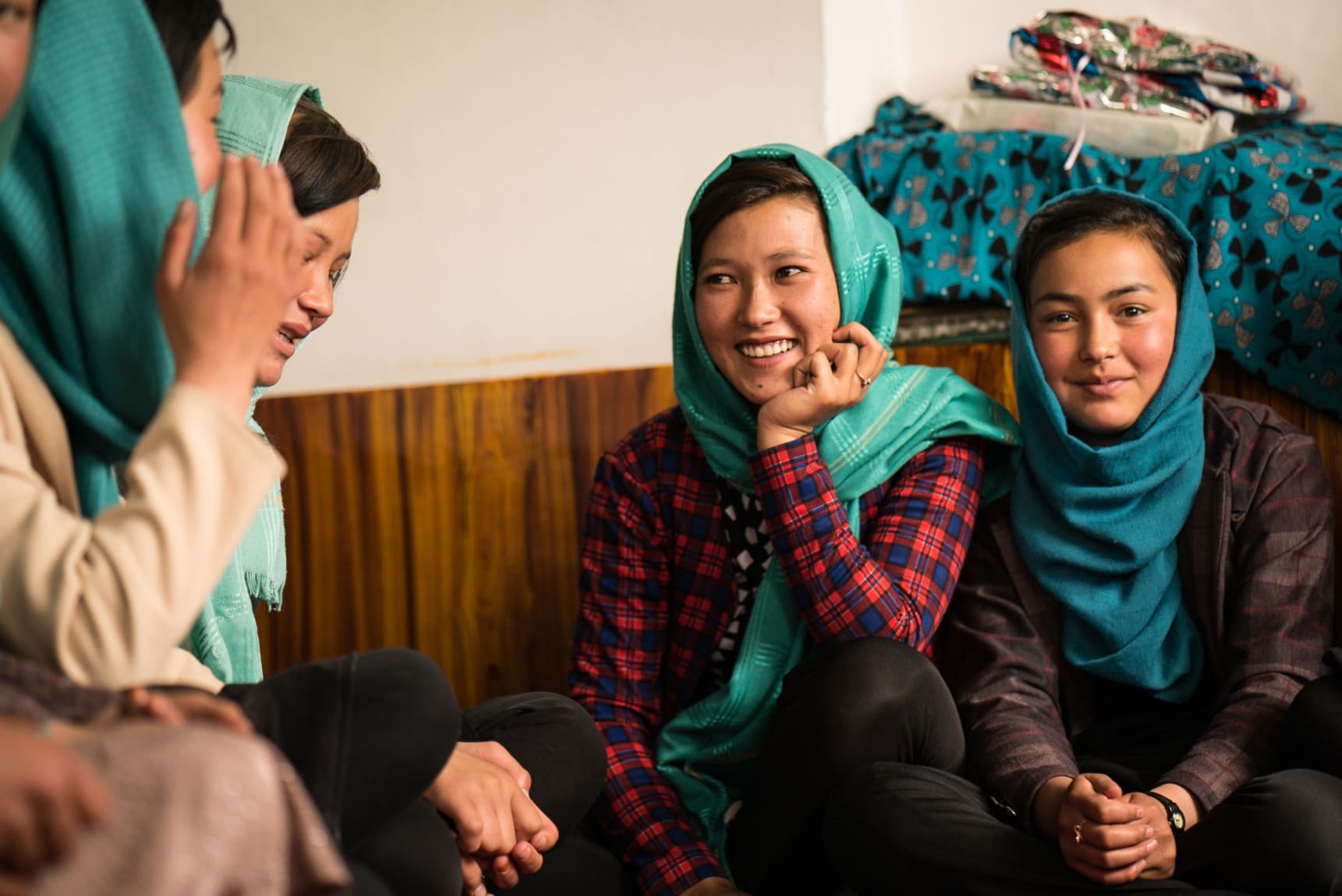 Against the backdrop of a white wall with a wood-effect sticker on the lower half, a girl wearing a purple plaid blazer, black trousers and teal hijab is pictured sitting cross-legged on the right, smiling and looking at the camera. Behind her is a piece of teal fabric draped over something and stacked with some unidentifiable shiny objects. On the left of the first girl is another wearing black trousers, a long-sleeved red and blue plaid top and turquoise hijab - she is smiling and resting her chin on her left hand as her left elbow is propped on her left knee. To the left of the frame we can see parts of the side profiles of 3 more women sat on the floor.
