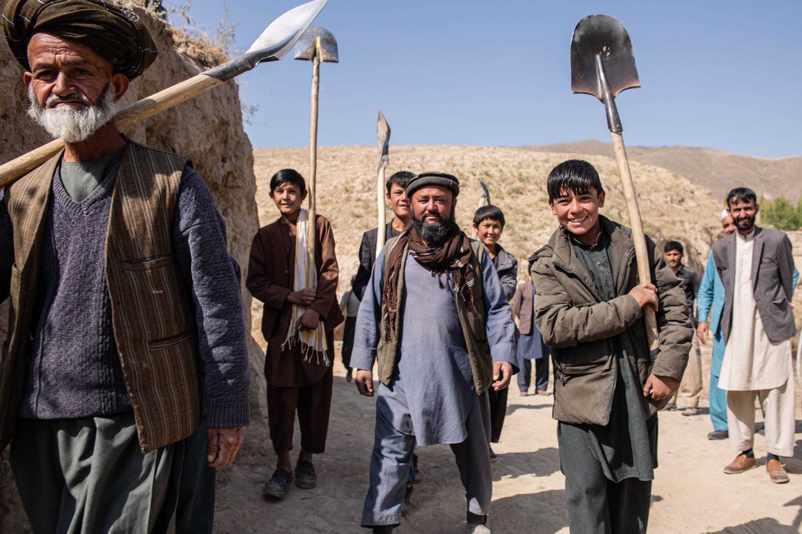 Several men are pictured against the backdrop of stone-coloured mountains under blue skies in Chil Kapa village in Badakhshan, Afghanistan, walking towards the camera. The young man at the front on the right is smiling wearing a dark brown puffer jac
